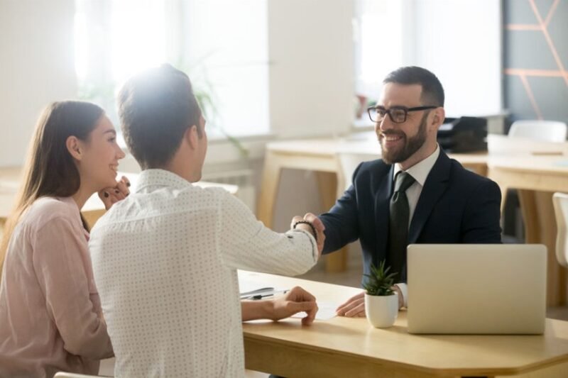 Un homme en costume serre la main d'un homme de l'autre côté d'un bureau, tandis qu'une femme est assise à côté d'eux, tous souriants dans un bureau.
