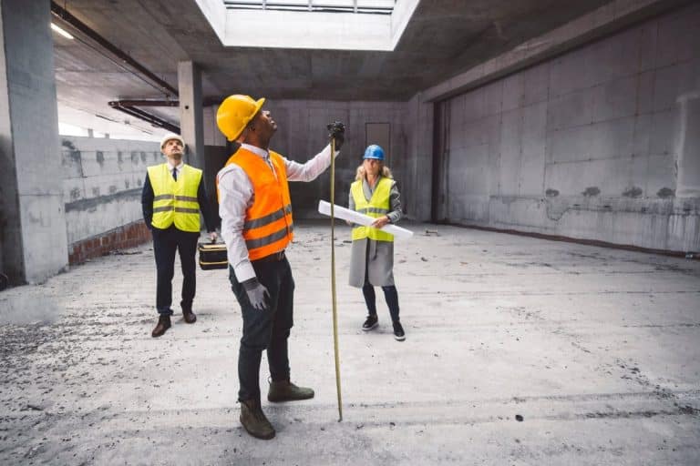 Trois ouvriers du bâtiment, équipés d'équipements de sécurité et munis d'outils, se tiennent dans un bâtiment inachevé. L'un mesure avec un mètre ruban tandis qu'un autre examine des plans.