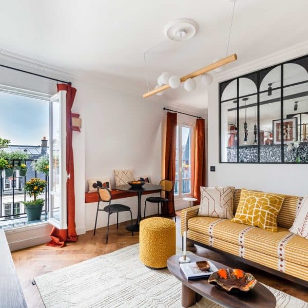 Séjour lumineux avec canapé jaune, portes fenêtres ouvertes, petite table à manger et miroir mural. Décoration comprenant des plantes en pot et des coussins à motifs. Plancher en bois et luminaires élégants.