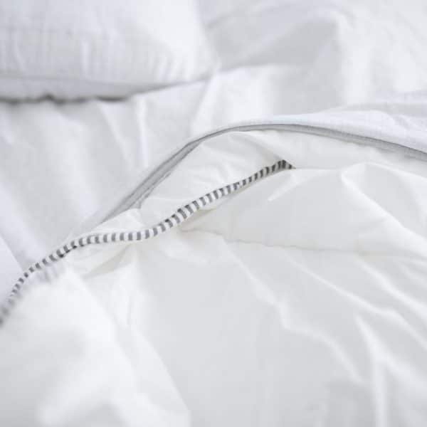 Gros plan d'un lit blanc avec une couette moelleuse à bordure rayée, repliée sur elle même. Des oreillers blancs en arrière plan.
