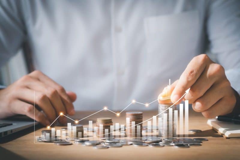 Une personne empile des pièces de monnaie sur une table à côté d'un ordinateur portable, avec une superposition numérique d'un graphique linéaire ascendant et d'un graphique à barres.