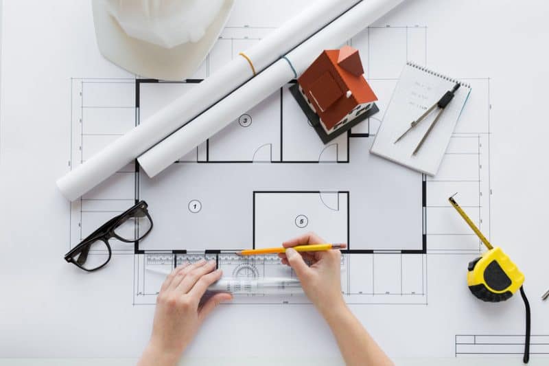Vue aérienne des mains utilisant une règle sur des plans architecturaux avec un casque, des lunettes et un ruban à mesurer sur une surface blanche.