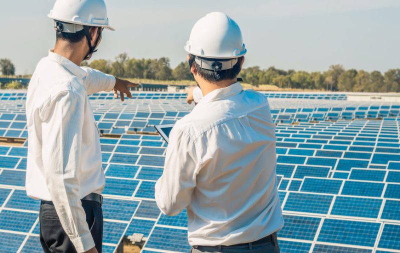 Deux ingénieurs portant des chemises blanches et des casques de chantier se tiennent devant des panneaux solaires, l'un pointant tandis que l'autre tient une tablette, dans une installation d'énergie solaire.
