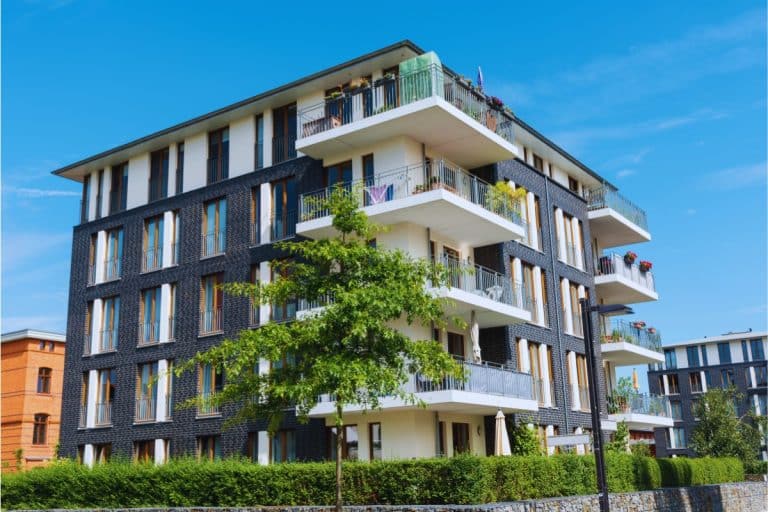 Immeuble résidentiel moderne à plusieurs étages avec balcons, entouré de verdure et d'un ciel bleu clair.