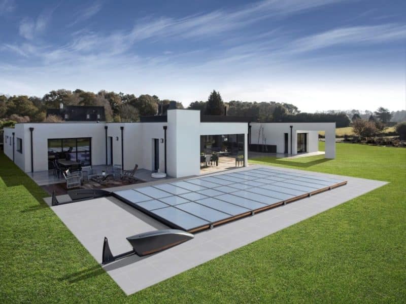Maison moderne blanche de plain pied avec de grandes fenêtres, un patio meublé et une piscine rectangulaire couverte, située sur une grande pelouse verte sous un ciel bleu.