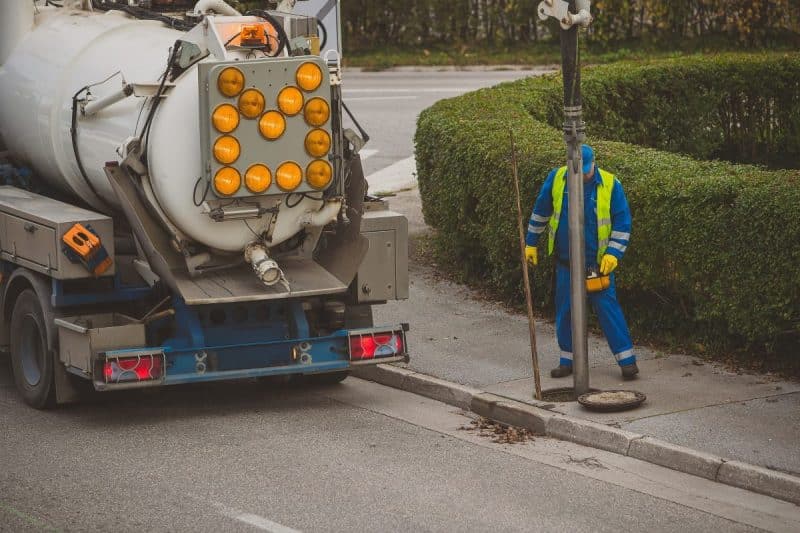 Un travailleur portant un équipement réfléchissant se tient près d'un camion utilitaire et d'un regard d'égout ouvert au coin d'une rue de banlieue.