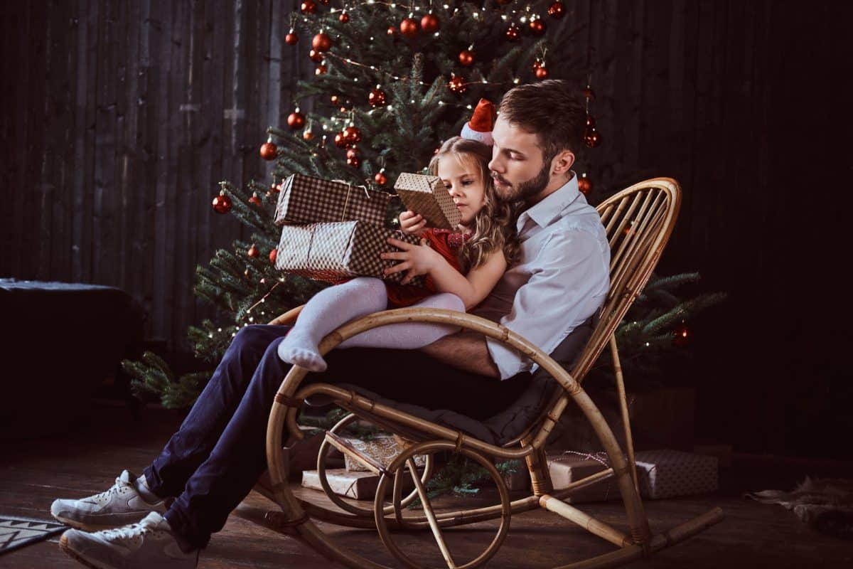 Un homme et un enfant assis dans un fauteuil à bascule devant un sapin de Noël décoré. L'enfant tient un cadeau emballé.