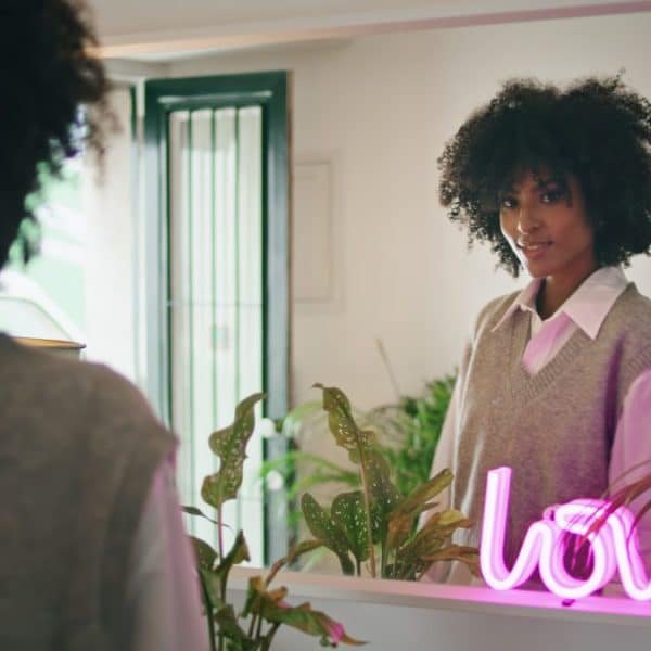 Une femme aux cheveux bouclés se regarde dans un miroir à côté de plantes et d'un néon rose qui dit "amour".