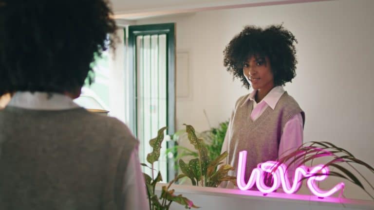 Une femme aux cheveux bouclés se regarde dans un miroir à côté de plantes et d'un néon rose qui dit "amour".