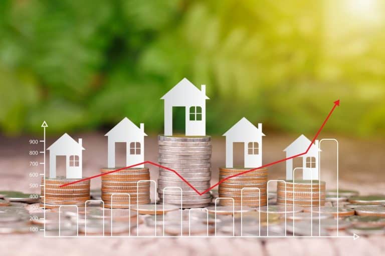 Des piles de pièces avec des icônes de maison sur le dessus, superposées à un graphique en ligne rouge, symbolisant les tendances du marché immobilier sur un fond vert flou.