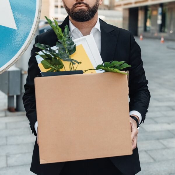 Un homme en costume tient une boîte en carton remplie de fournitures de bureau et de plantes, debout sur un trottoir de la ville.