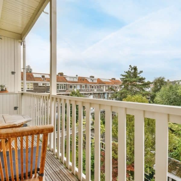 Un balcon en bois avec des chaises et une petite table donne sur un quartier résidentiel et des arbres sous un ciel partiellement nuageux.