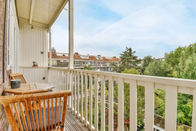 Un balcon en bois avec des chaises et une petite table donne sur un quartier résidentiel et des arbres sous un ciel partiellement nuageux.