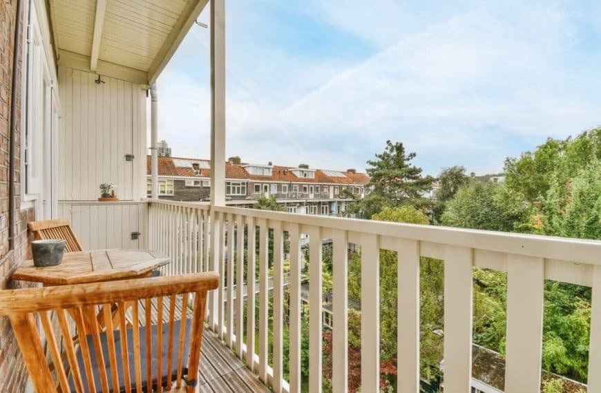 Un balcon en bois avec des chaises et une petite table donne sur un quartier résidentiel et des arbres sous un ciel partiellement nuageux.