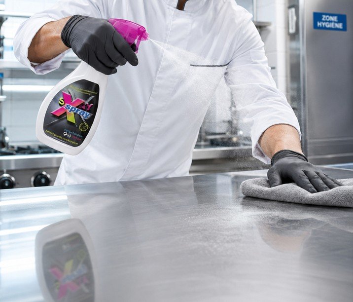 Dans une cuisine professionnelle, une personne vêtue de gants noirs et d'une blouse blanche vaporise un produit nettoyant sur une surface en acier inoxydable et l'essuie avec un chiffon.