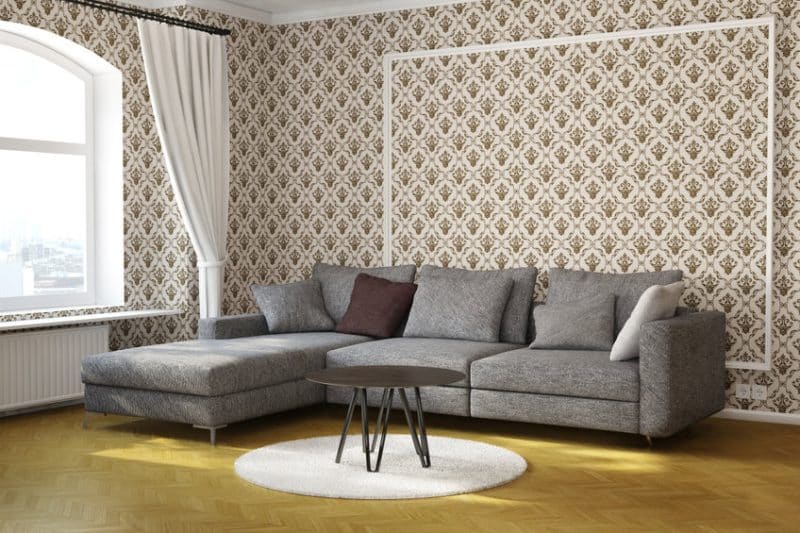 Un canapé sectionnel gris avec des coussins et un coussin marron se trouve dans un salon avec du papier peint à motifs et une table basse ronde sur un tapis blanc.
