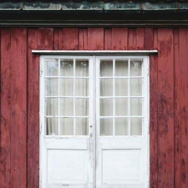 Mur en bois rouge avec une double porte blanche patinée et une rangée de petites fenêtres carrées.