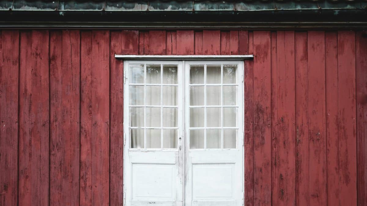 Mur en bois rouge avec une double porte blanche patinée et une rangée de petites fenêtres carrées.