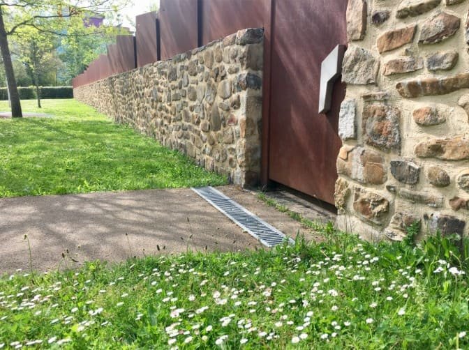 Un mur de pierre longe une pelouse parsemée de marguerites, à côté d'un chemin pavé et d'une grille métallique enfoncée dans le sol.