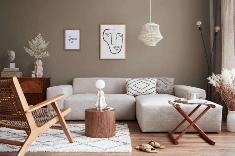 Un salon contemporain avec un canapé modulable gris clair, un fauteuil en bois, une table basse ronde en bois et un tapis blanc. Des œuvres d'art murales, une suspension en osier et des plantes séchées rehaussent le décor.