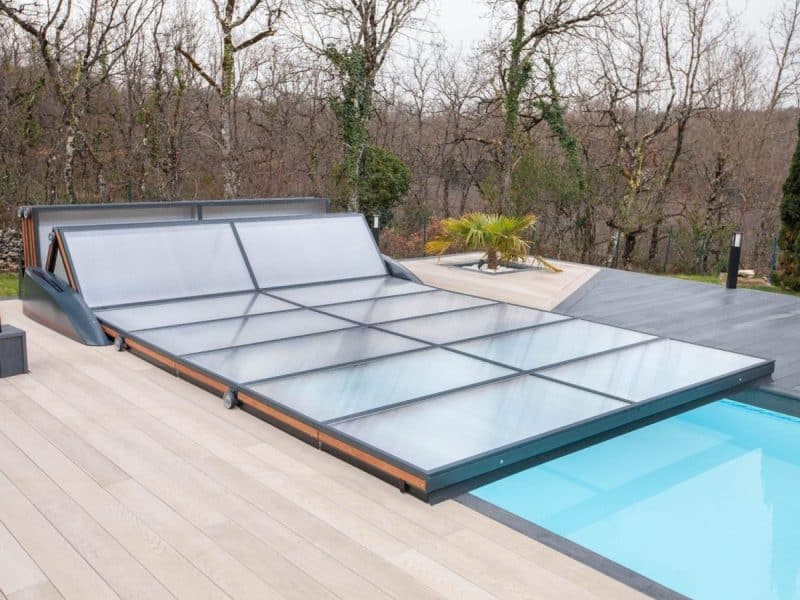 Une couverture de piscine rétractable partiellement étendue sur une piscine extérieure, entourée d'une terrasse et d'arbres en arrière plan.