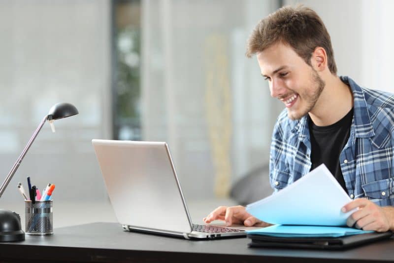 Jeune homme souriant et travaillant sur un ordinateur portable à un bureau avec des documents et des fournitures de bureau.