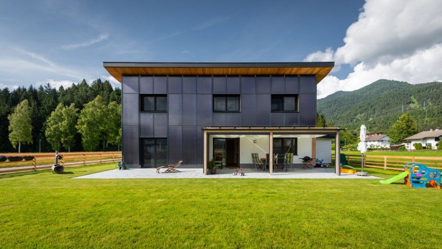Une maison moderne à deux étages avec des panneaux extérieurs foncés et un patio couvert, située sur une grande cour herbeuse avec des équipements de jeux pour enfants, entourée de montagnes et d'arbres.