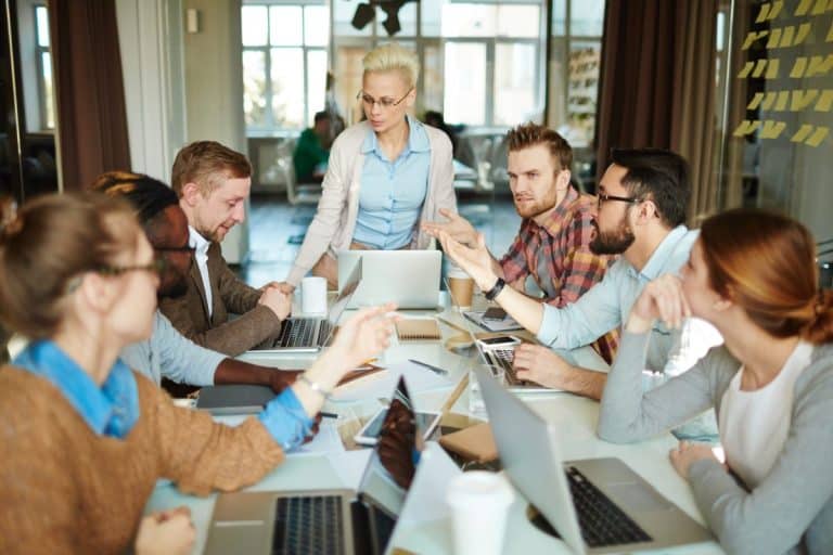 Un groupe de personnes dans une salle de réunion, assises autour d'une table avec des ordinateurs portables et des documents, discutent. Une personne, debout, observe et écoute la conversation.