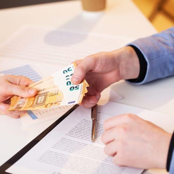 Une personne remet une pile de billets de banque en euros à une autre personne qui signe un document sur un bureau.