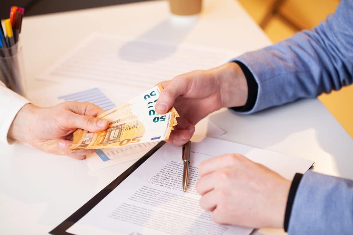 Une personne remet une pile de billets de banque en euros à une autre personne qui signe un document sur un bureau.