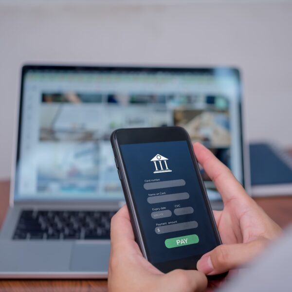 Une personne tient un smartphone affichant un formulaire de paiement en ligne, avec un ordinateur portable ouvert à l'arrière-plan sur un bureau.