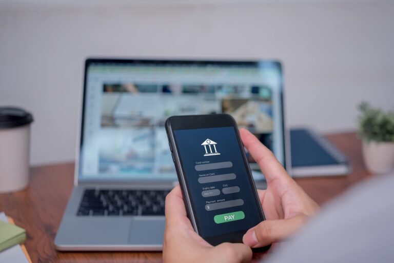 Une personne tient un smartphone affichant un formulaire de paiement en ligne, avec un ordinateur portable ouvert à l'arrière-plan sur un bureau.