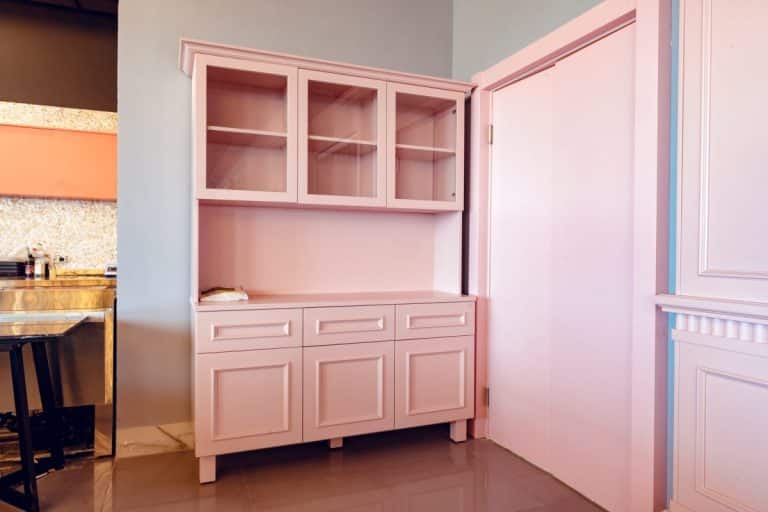 Ensemble d'armoires de cuisine roses avec portes supérieures en verre et tiroirs inférieurs solides, placés contre un mur rose clair.