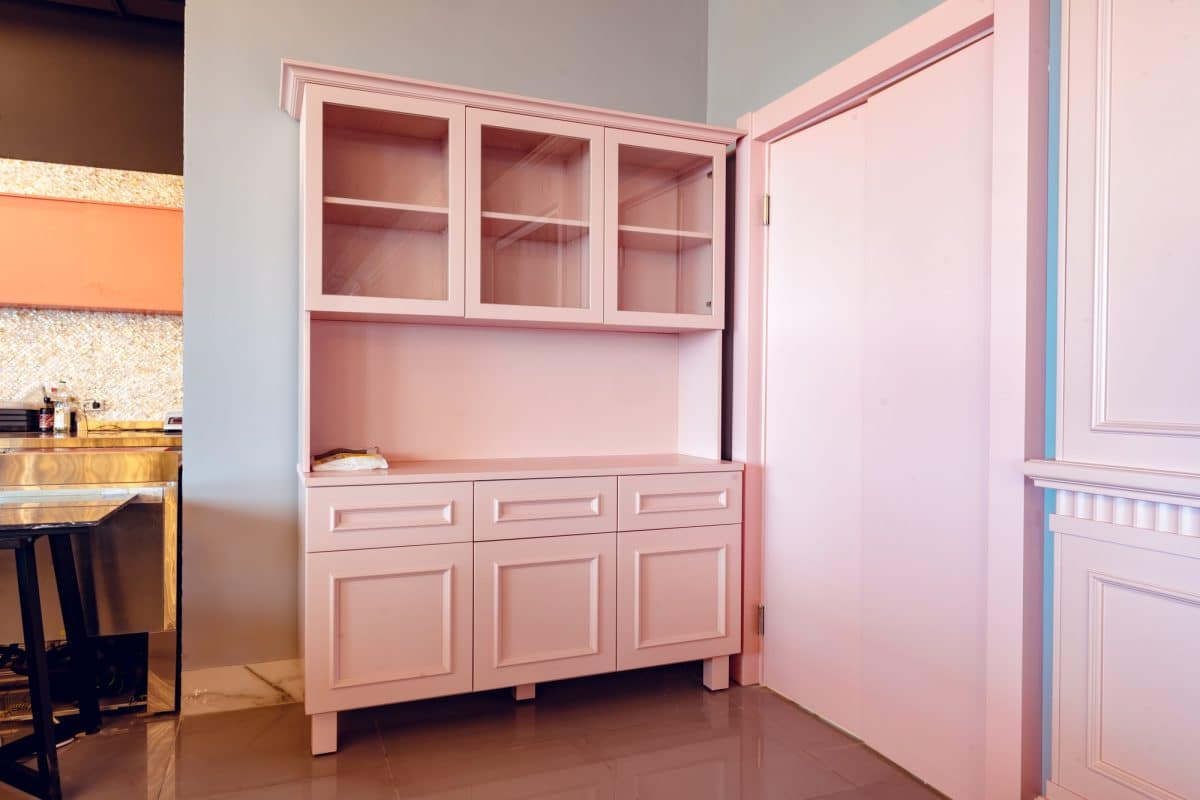Ensemble d'armoires de cuisine roses avec portes supérieures en verre et tiroirs inférieurs solides, placés contre un mur rose clair.