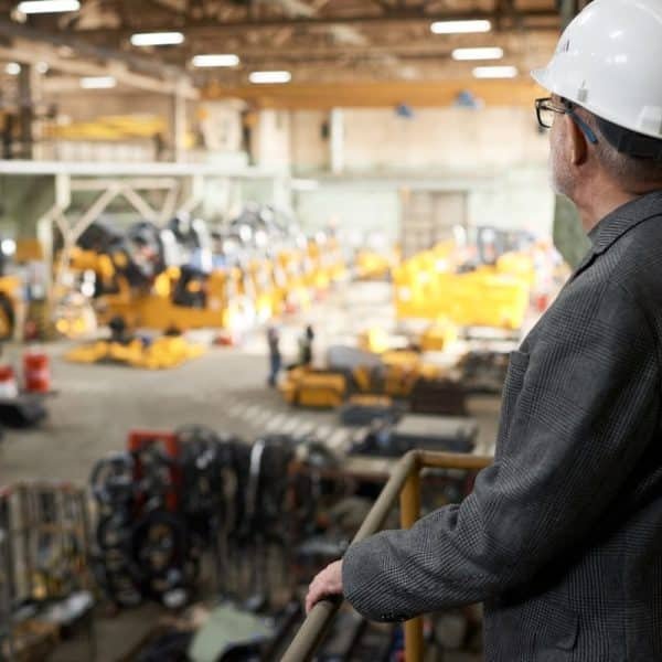 Un homme portant un casque et un costume observe les machines et l'équipement à l'intérieur d'un grand entrepôt industriel depuis une position élevée.