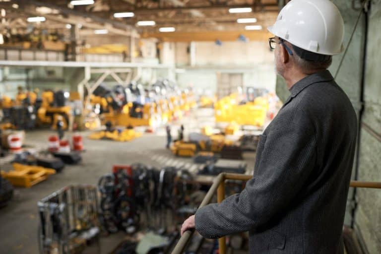Un homme portant un casque et un costume observe les machines et l'équipement à l'intérieur d'un grand entrepôt industriel depuis une position élevée.