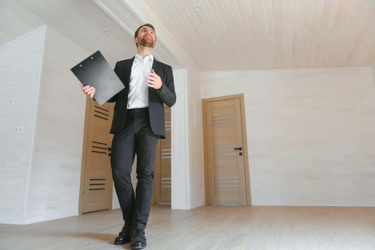 Un homme en costume tient un presse papiers tout en marchant dans une pièce non meublée avec du parquet et des murs blancs.