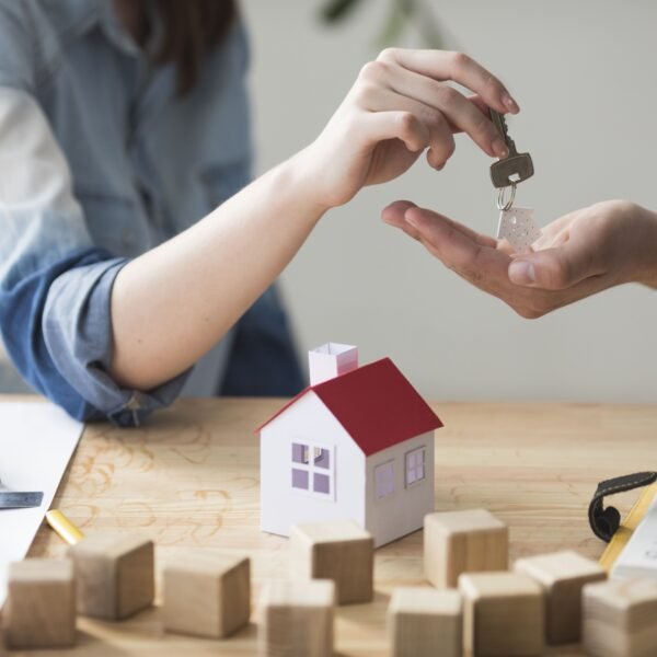 Une personne remet les clés d'une maison à une autre au dessus d'une table sur laquelle se trouvent des documents, une petite maquette de maison et des cubes en bois.