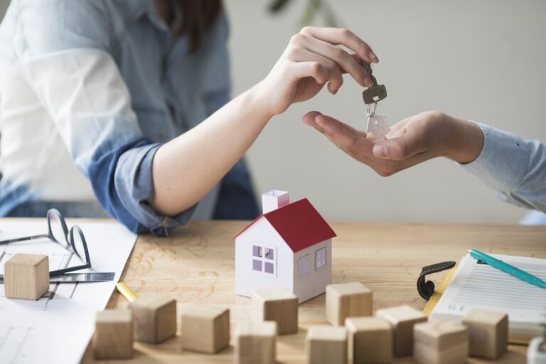 Une personne remet les clés d'une maison à une autre au dessus d'une table sur laquelle se trouvent des documents, une petite maquette de maison et des cubes en bois.