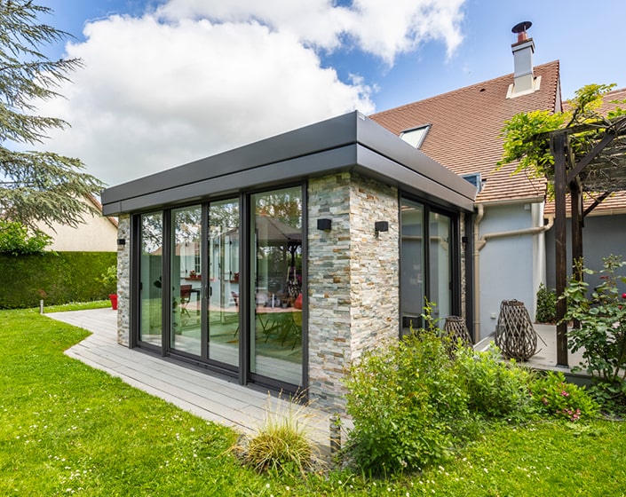 Maison moderne avec une extension aux murs vitrés, des accents de pierre et un toit en pente. Entourée d'une pelouse verte luxuriante et d'arbustes sous un ciel partiellement nuageux.