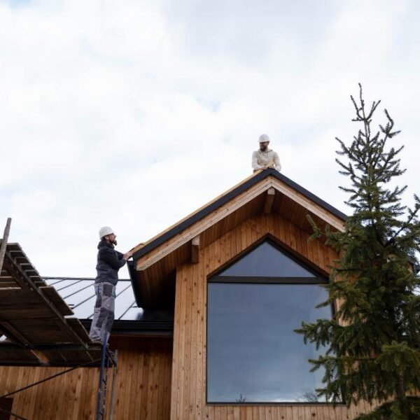 Deux ouvriers du bâtiment portant des casques de protection travaillent sur le toit d'une maison en bois ; l'un est sur un échafaudage tandis que l'autre est sur le faîte du toit.