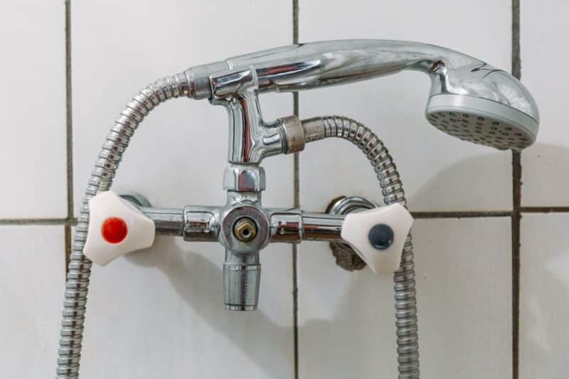 Gros plan d'un robinet de douche avec tuyaux métalliques et boutons d'eau chaude et froide séparés devant un mur carrelé.
