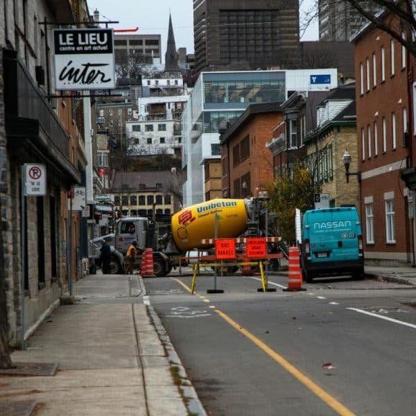 Une rue de la ville est bloquée par des barrières de construction, un camion de ciment et une camionnette ; des bâtiments bordent les deux côtés et des panneaux de signalisation sont visibles.