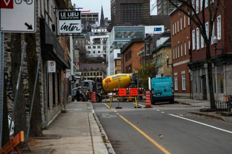 Une rue de la ville est bloquée par des barrières de construction, un camion de ciment et une camionnette ; des bâtiments bordent les deux côtés et des panneaux de signalisation sont visibles.