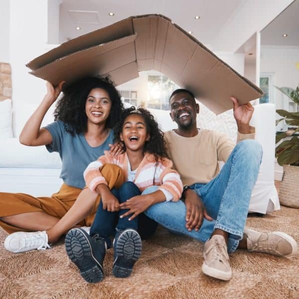Une famille souriante de trois personnes est assise par terre dans un salon, tenant un grand morceau de carton en forme de toit au dessus de leur tête.