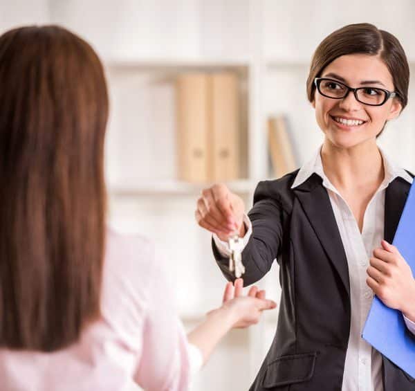 Une femme d'affaires en costume remet les clés à une autre personne. Elle tient un dossier bleu et sourit.
