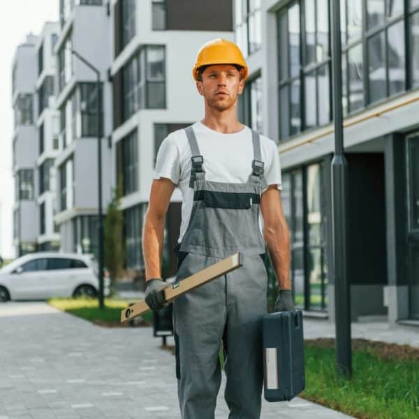 Un ouvrier du bâtiment portant un casque de sécurité et une salopette se tient sur un chemin pavé entre des immeubles d'appartements modernes, tenant une boîte à outils et un outil de mesure.