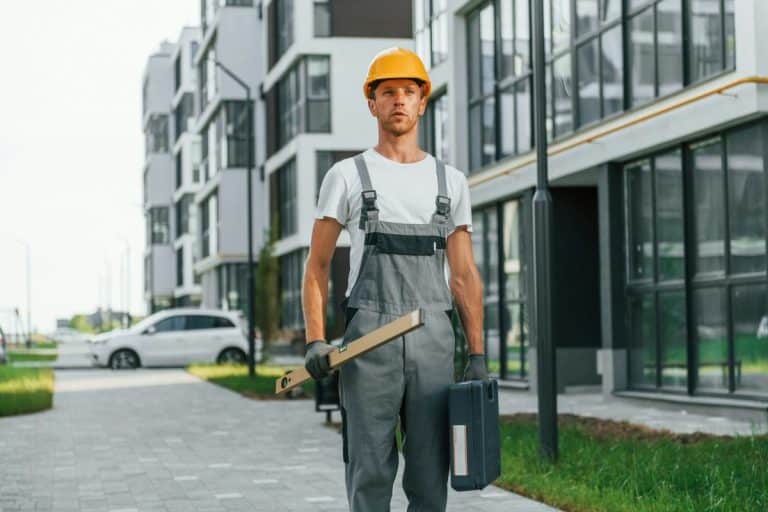 Un ouvrier du bâtiment portant un casque de sécurité et une salopette se tient sur un chemin pavé entre des immeubles d'appartements modernes, tenant une boîte à outils et un outil de mesure.