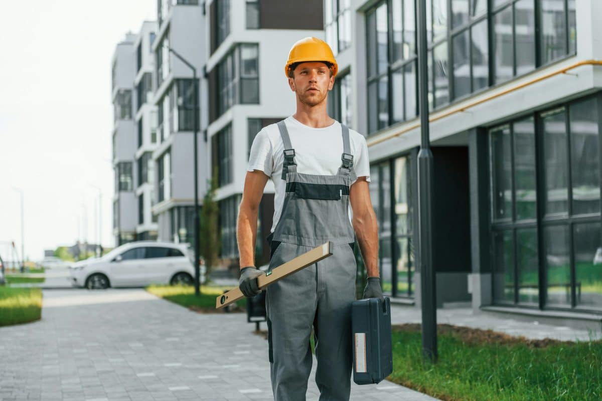 Un ouvrier du bâtiment portant un casque de sécurité et une salopette se tient sur un chemin pavé entre des immeubles d'appartements modernes, tenant une boîte à outils et un outil de mesure.