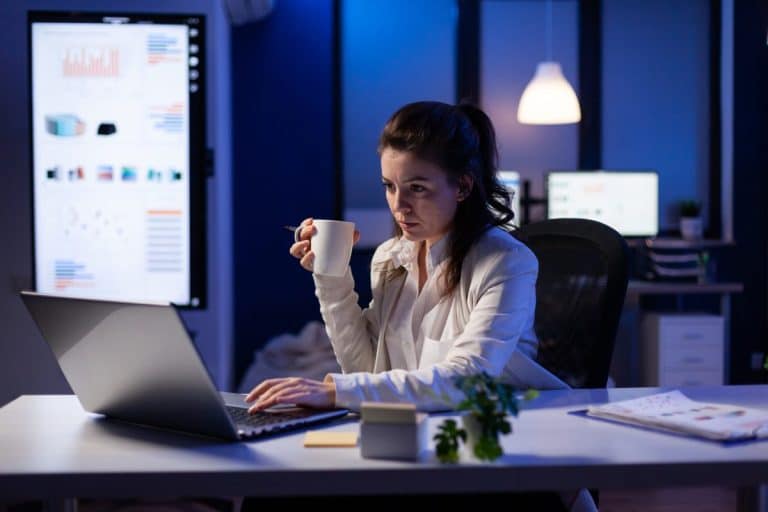 Une femme est assise à un bureau, travaillant sur un ordinateur portable la nuit, tenant une tasse, avec des graphiques affichés sur des écrans en arrière plan.